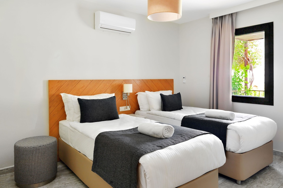 A hotel room with two beds, a wooden headboard, a bedside lamp, and a window with a view of greenery.