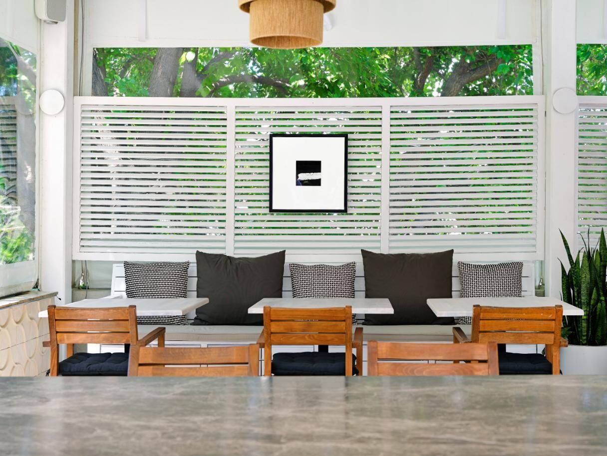 A modern outdoor seating area with a sofa and wooden chairs, a framed picture on the wall, and a view of trees through the window blinds.