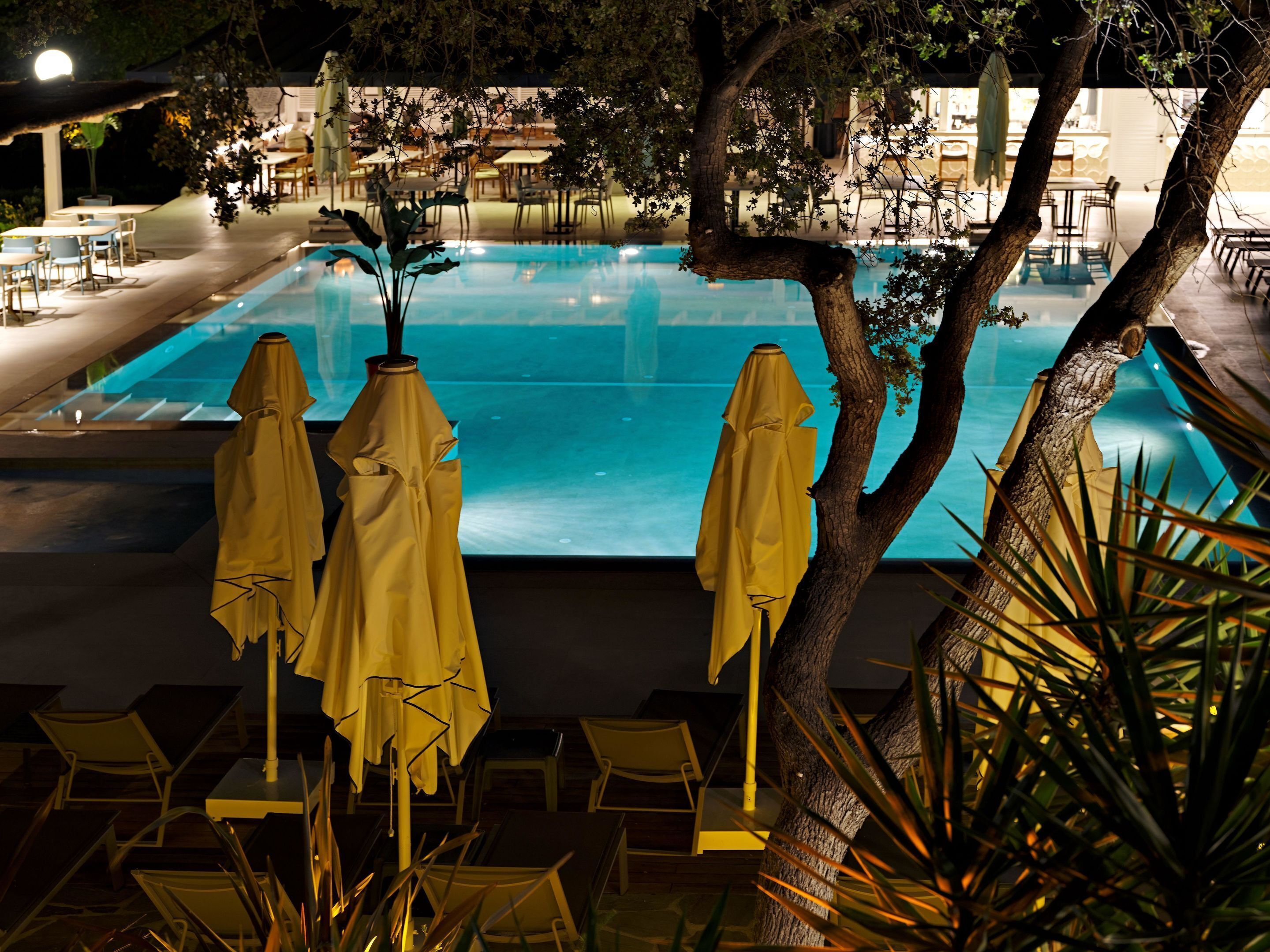 A pool area with lounge chairs and umbrellas, surrounded by trees and a building in the background.