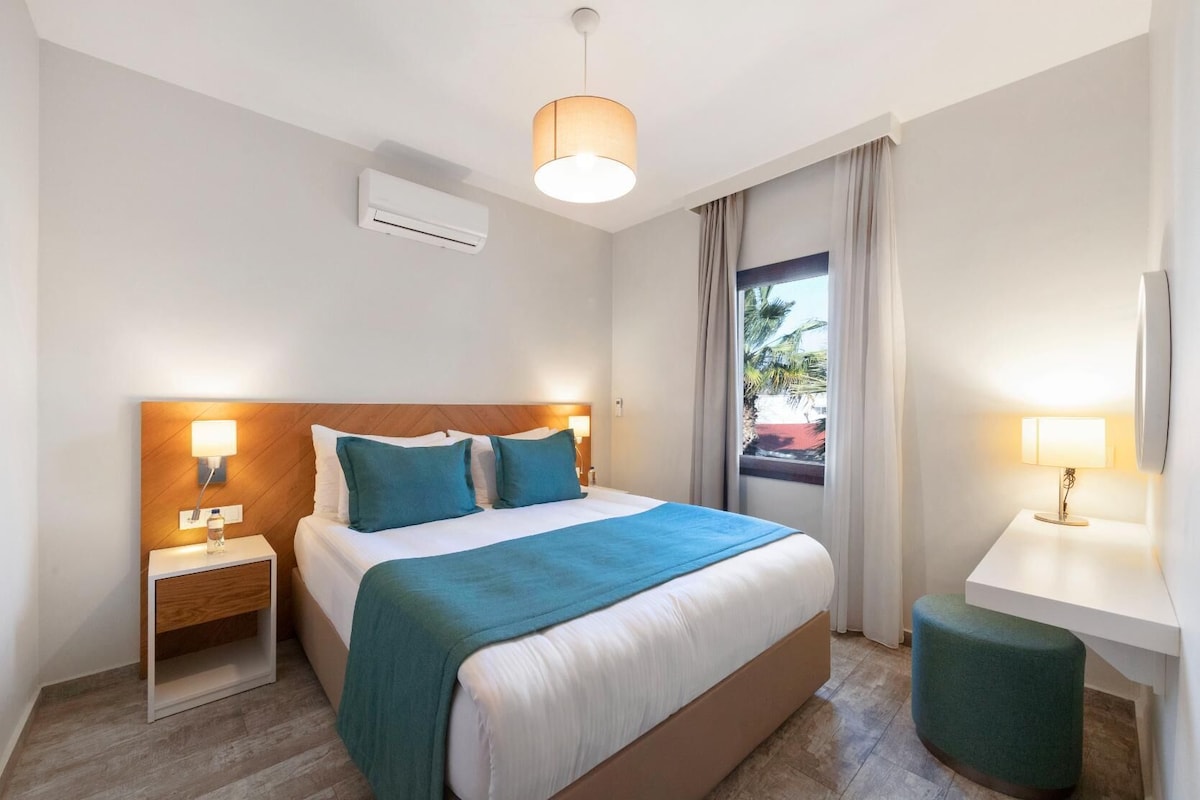 A hotel room with a bed, bedside tables, a nightstand, a desk, a chair, an air conditioning unit, and a window with curtains.
