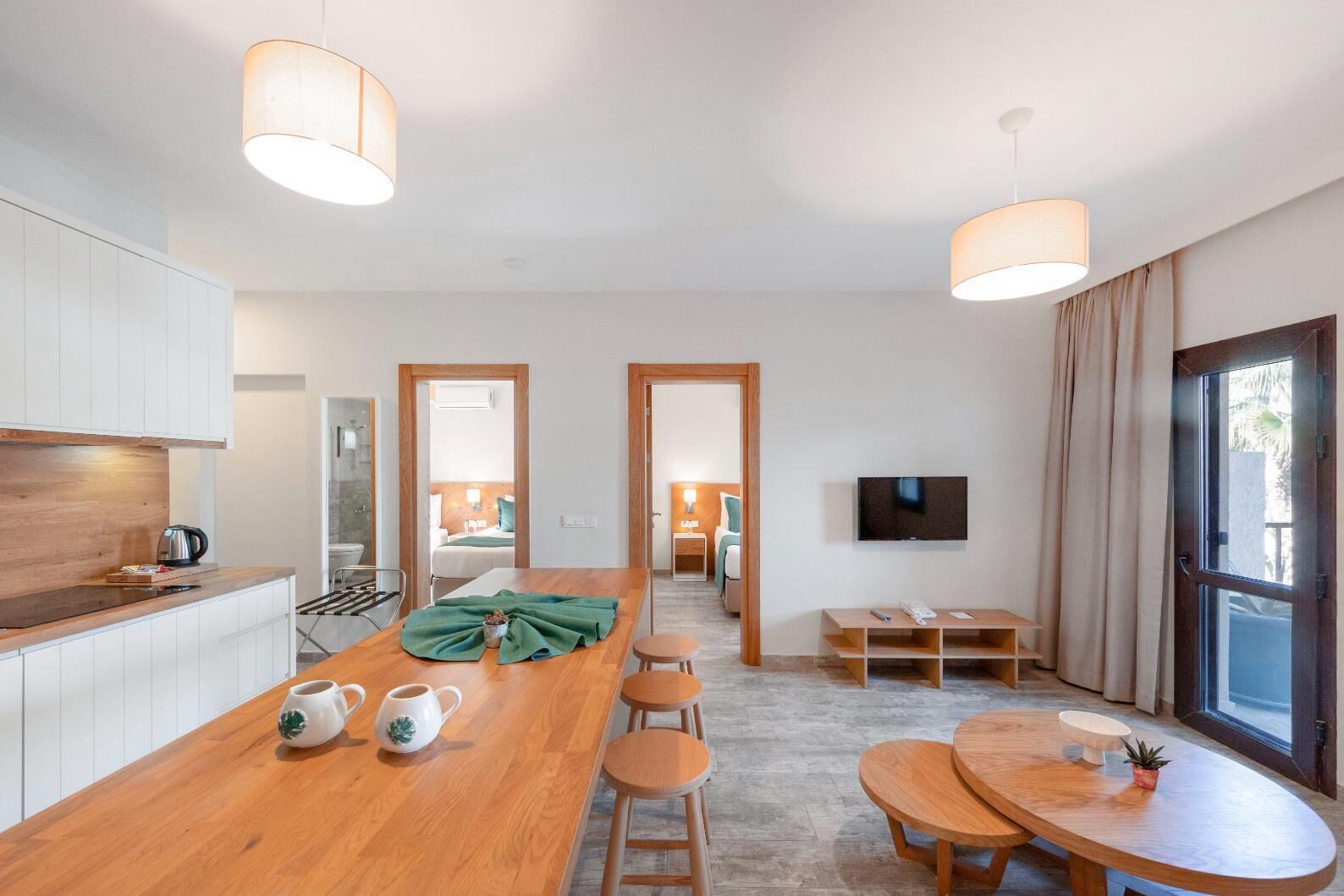 A modern hotel room with a wooden dining table, bar stools, a kitchen area, a TV, and a large window with curtains.