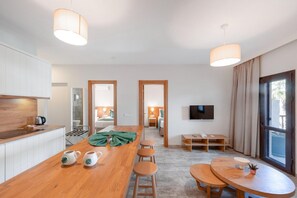 Una habitación de hotel moderna con una mesa de comedor de madera, taburetes de bar, una zona de cocina, un televisor y un ventanal con cortinas.