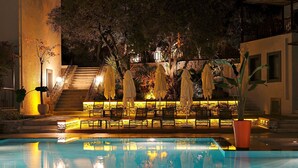 Un espace piscine éclairé la nuit, avec des chaises longues et des parasols, un escalier en pierre et un bâtiment en arrière-plan.