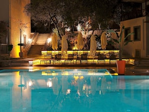 A nighttime pool area with lounge chairs and umbrellas, a stone staircase, and a building in the background.