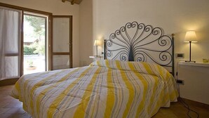 Quarto com cama, cabeceira metálica decorativa, mesa de cabeceira com abajur e vista para o exterior através de uma porta.