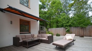 Un espace extérieur aménagé, abrité, avec un canapé et un fauteuil de relaxation, une terrasse en bois et une clôture en bois en arrière-plan.