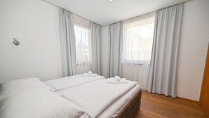 Une chambre double avec deux lits, des rideaux clairs, un plancher en bois et une petite lampe fixée au mur.
