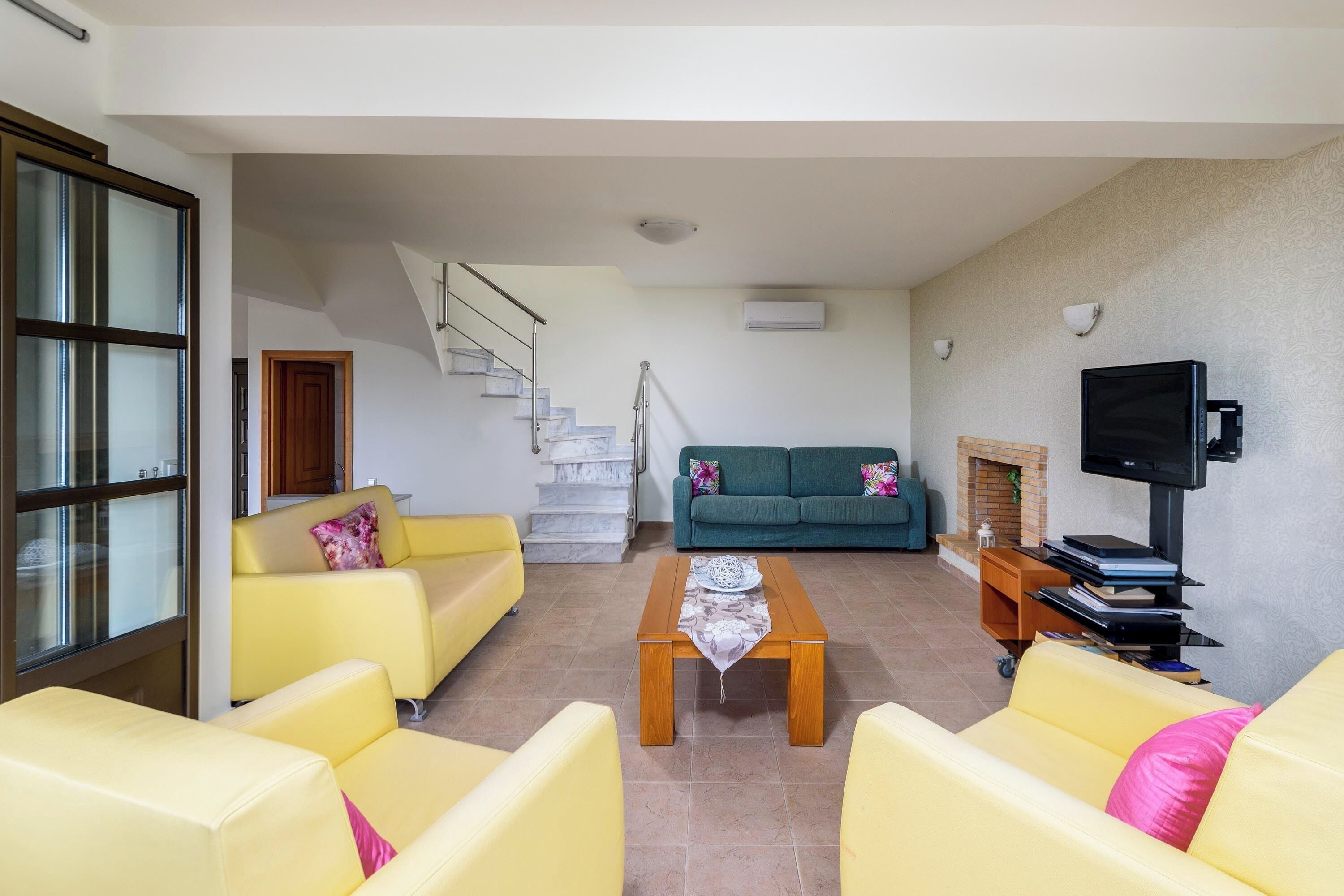 Una sala de estar con sofás amarillos, una mesa de centro de madera, un sofá verde, un televisor y una escalera que conduce a otra habitación.