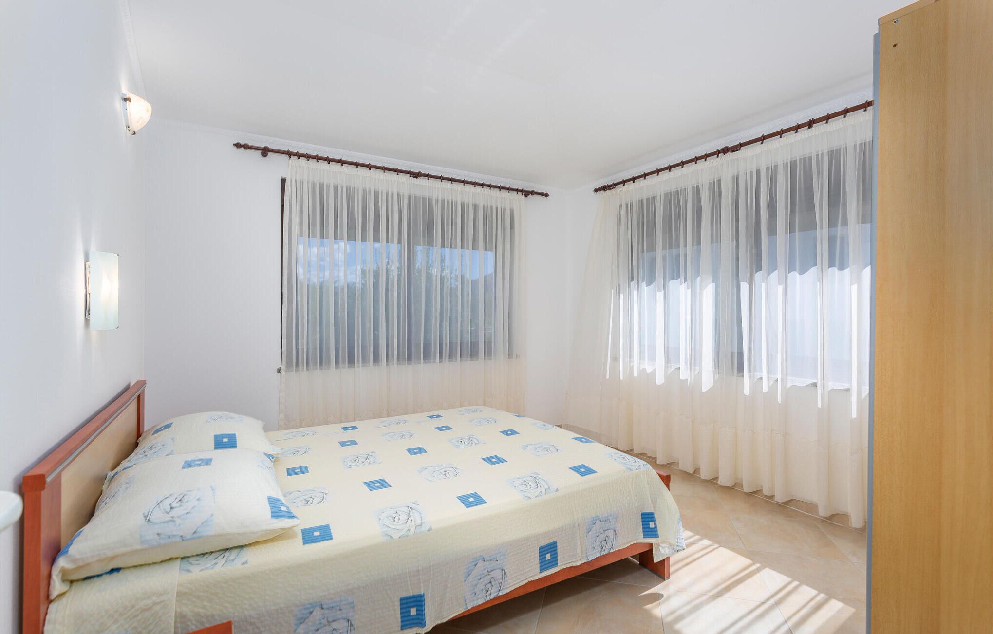 A bedroom with a bed, two windows with sheer curtains, a wooden headboard, and a light fixture on the wall.