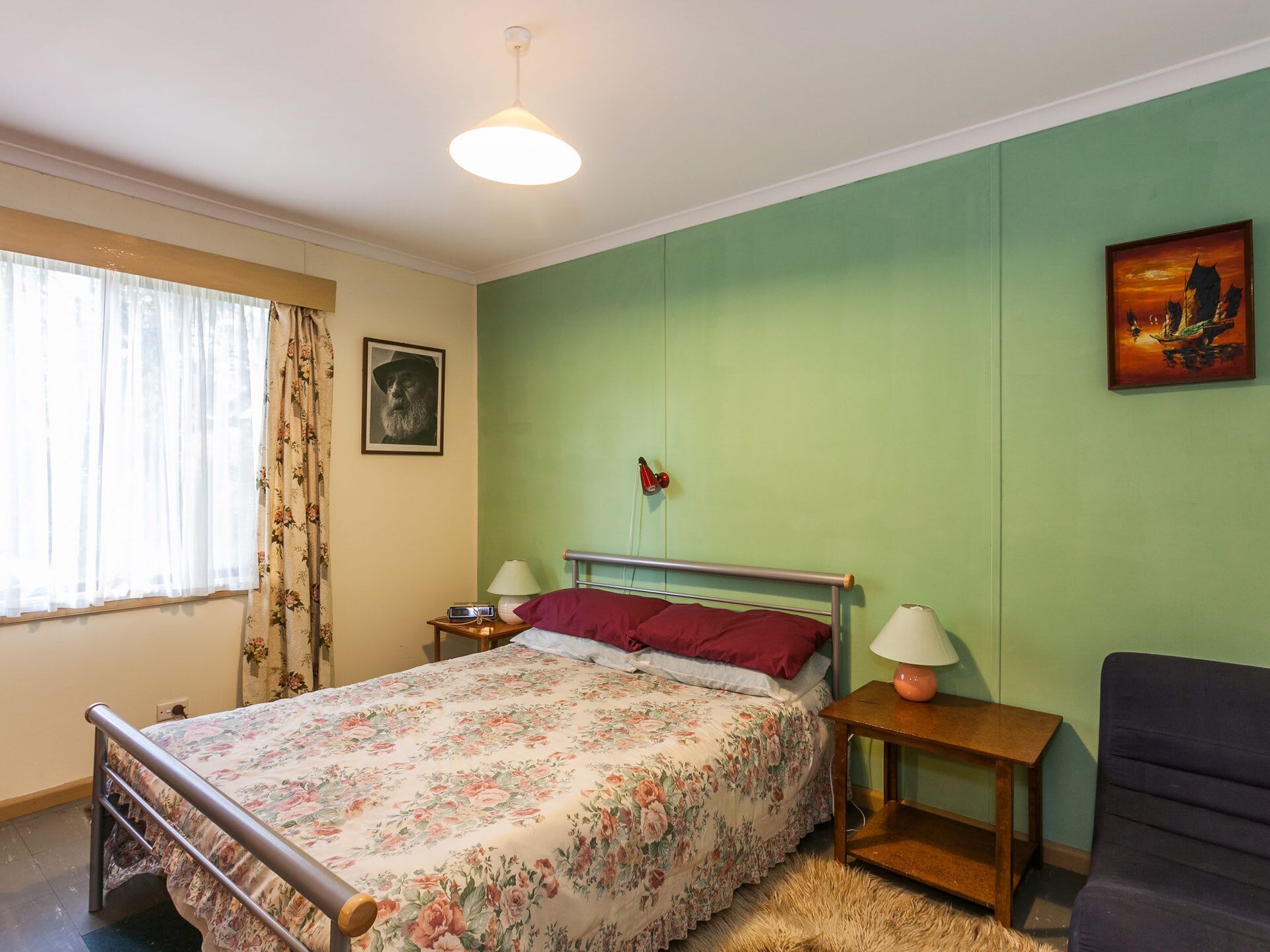Une chambre à coucher comprenant un lit, une table de chevet, une lampe, une chaise, un tableau et une fenêtre avec des rideaux.