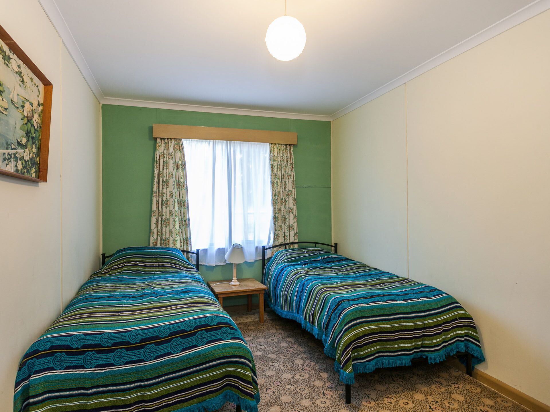 A room with two single beds, a window with curtains, a lamp, and a painting on the wall.