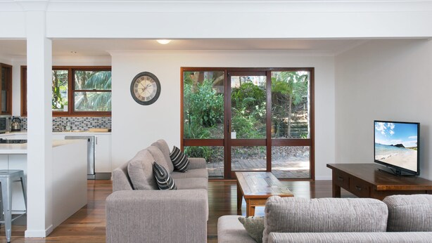 A spacious living area with a sofa, a coffee table, and a television. There is a kitchen area with a refrigerator and a window overlooking a garden. - Barefoot Beach House Shoal Bay Retreat (Shoal Bay)
