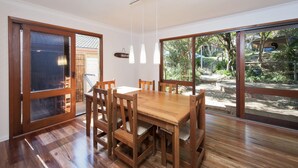 A dining area with a wooden table and chairs, large windows, and a view of a garden. - Barefoot Beach House Shoal Bay Retreat (Shoal Bay)