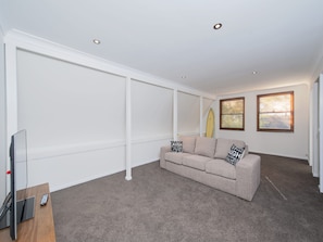 A living room with a beige sofa, a flat-screen TV, and a surfboard against the wall. - Barefoot Beach House Shoal Bay Retreat (Shoal Bay)