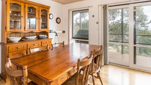 Een eethoek met een houten tafel en stoelen, een houten kast met glazen deuren en een groot raam met uitzicht op bomen.