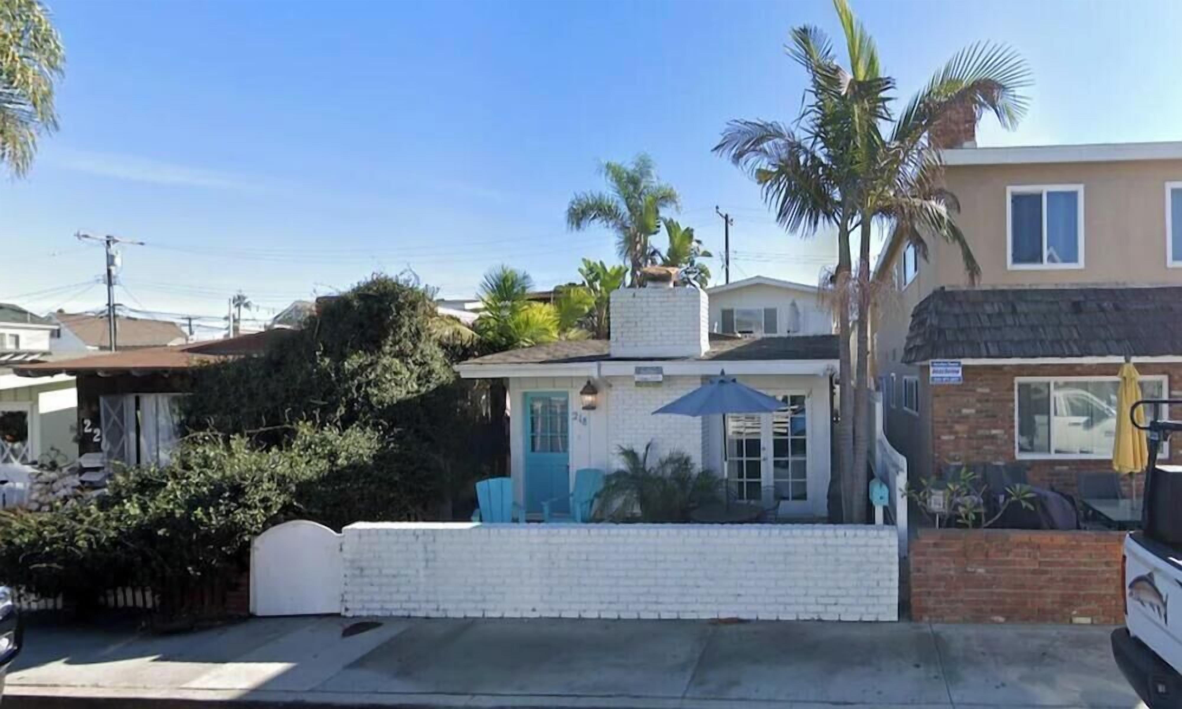 A small, single-story building with a white exterior and a blue door, surrounded by greenery and a clear sky.