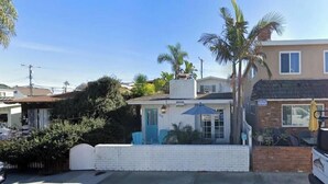 A small, single-story building with a white exterior and a blue door, surrounded by greenery and a clear sky. - Charming Beach Cottage Duplex-Huge Upper Level  (Newport Beach)