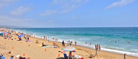 Een druk strand met mensen die zonnebaden en zwemmen, en een helder blauwe lucht.