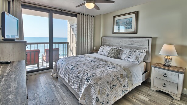 Une chambre à coucher comprenant un lit, une table de chevet, un téléviseur, un ventilateur de plafond et un balcon avec des chaises rouges.