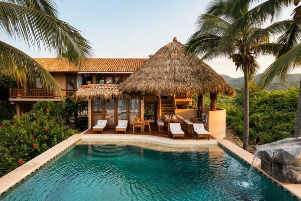 Un espace aménagé au bord de la piscine, avec des chaises longues, une structure à toit de chaume et une vue sur un bâtiment dont le toit est recouvert de tuiles.