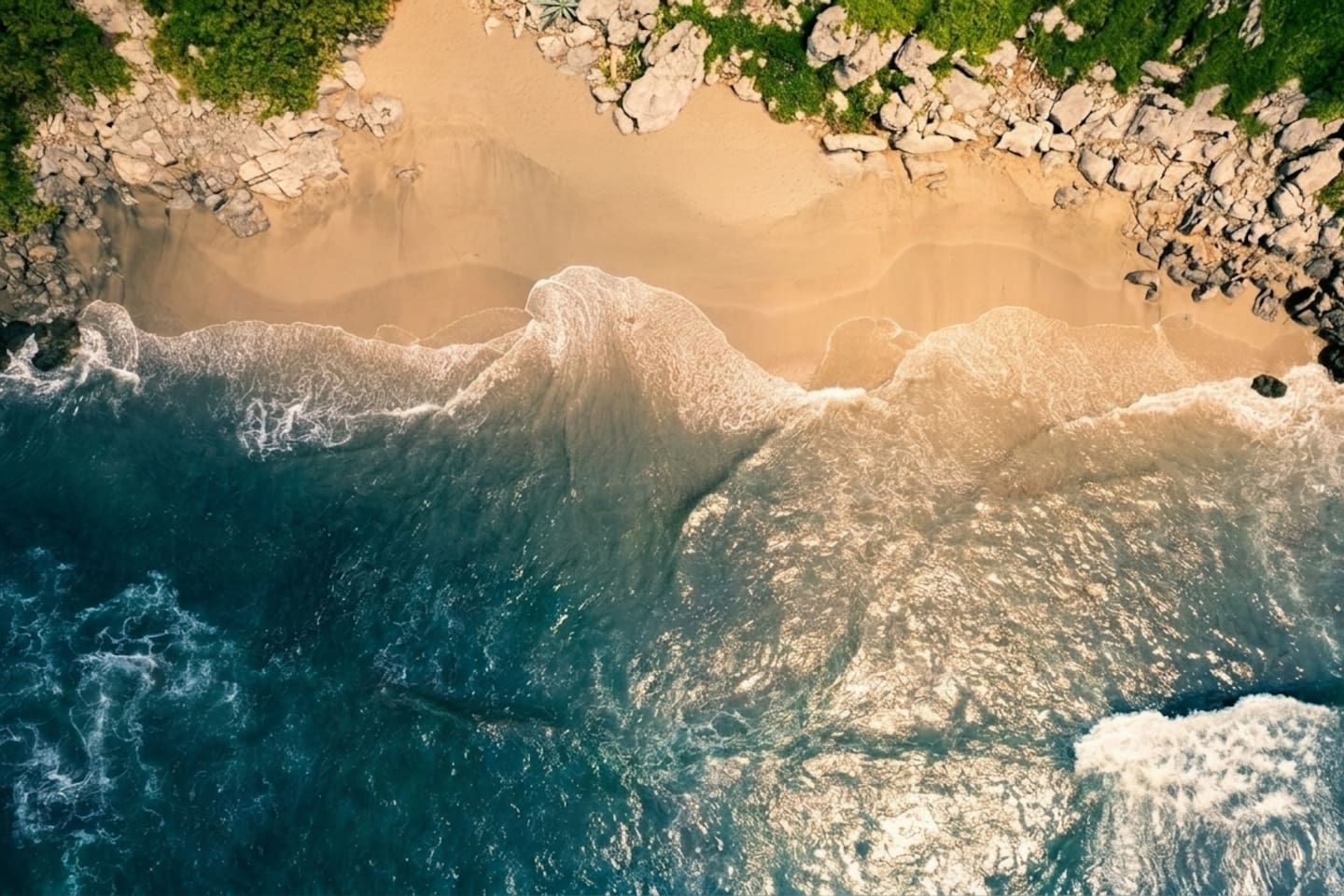 An aerial view of a beach with waves crashing onto the shore, surrounded by rocky terrain and sparse vegetation.