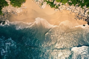 An aerial view of a beach with waves crashing onto the shore, surrounded by rocky terrain and sparse vegetation.