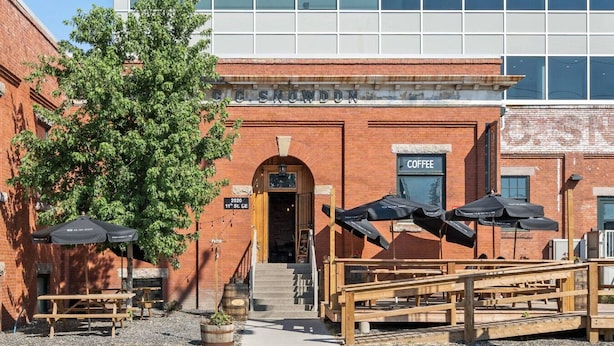 A brick building with a coffee shop sign, outdoor seating with umbrellas, and a staircase leading to the entrance. - Ramsay Manor | Spacious 1BR Queen Bed Inner City (Calgary)
