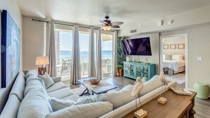 Ein geräumiges Wohnzimmer mit einer großen Couch, einem Fernseher und Meerblick durch große Fenster.