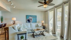 Ein geräumiges Wohnzimmer mit einem weißen Sofa, einem Couchtisch und einem Deckenventilator. An der Wand hängt ein Vogelgemälde, und durch die Fenster ist ein Balkon mit Gartenmöbeln zu sehen.