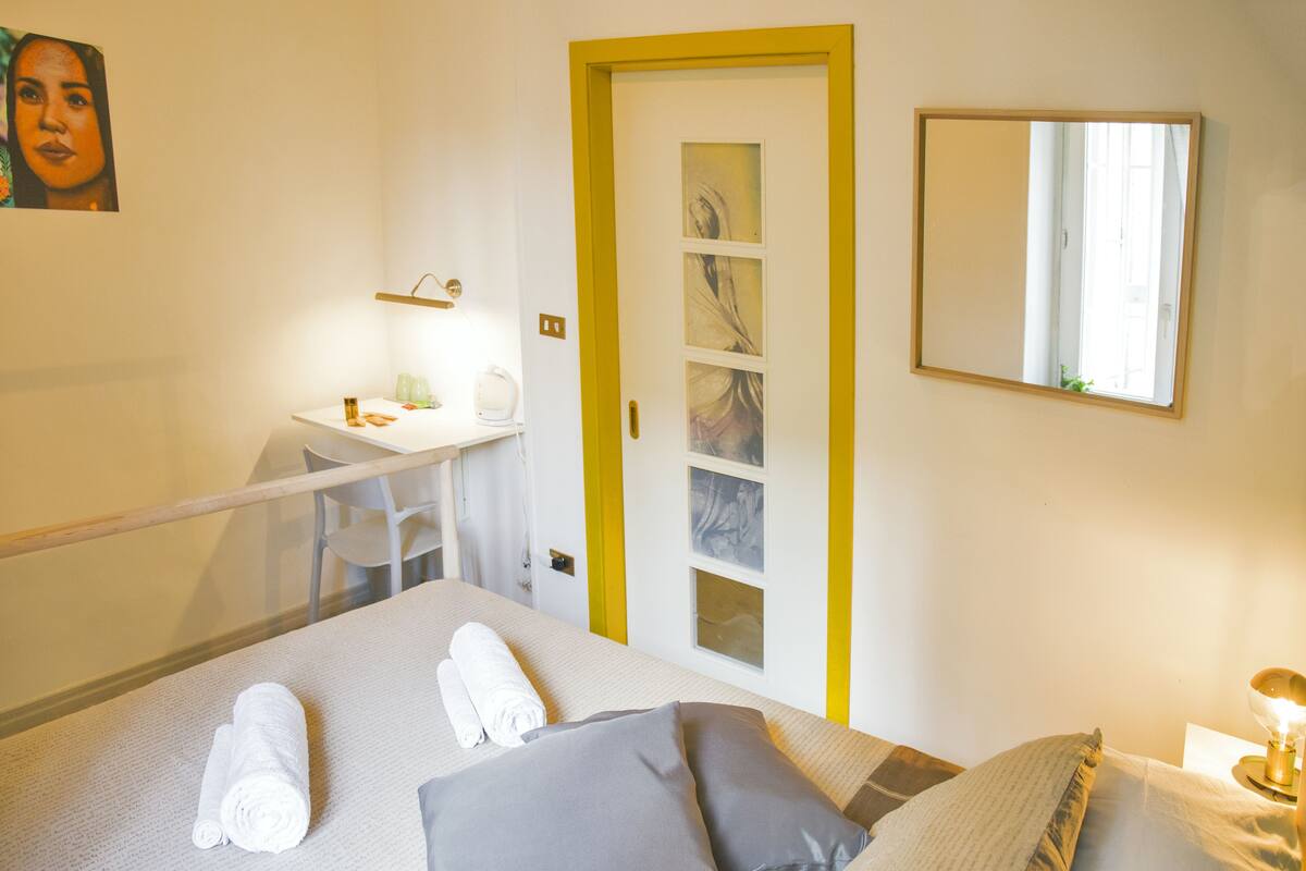 Habitación de hotel con cama, escritorio con silla, espejo y puerta con marco amarillo.