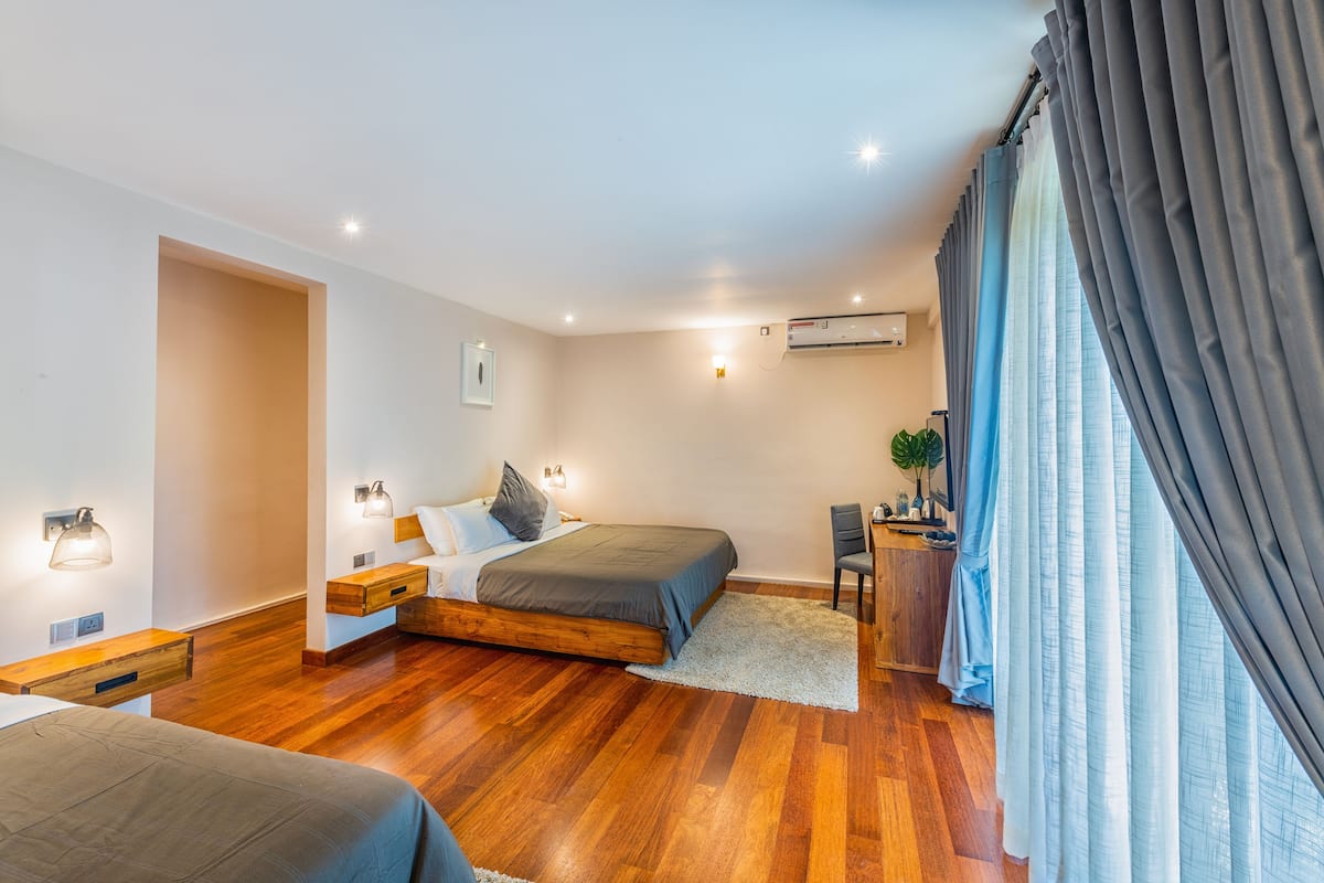 A modern bedroom with a wooden floor, a bed with a grey headboard, a desk with a chair, and a window with curtains.