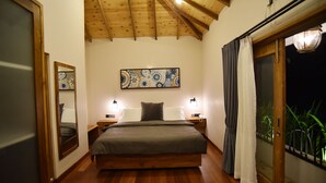 Un dormitorio con techo de madera, una cama con colcha gris, dos lámparas de noche, una obra de arte abstracto en la pared y una puerta corrediza de vidrio con cortinas.