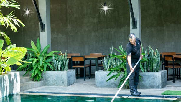 Zona de piscina con una persona limpiándola, rodeada de plantas en macetas y mobiliario de exterior.