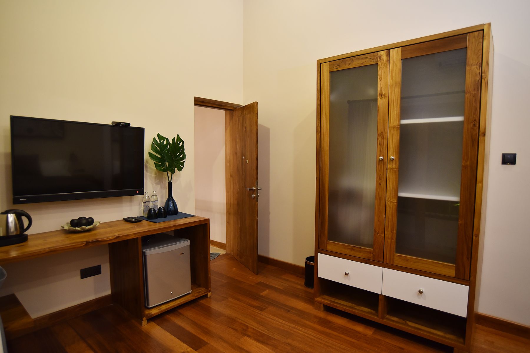 Una habitación con un escritorio de madera, un televisor, un refrigerador pequeño, un mueble con puertas de vidrio y una planta en maceta.