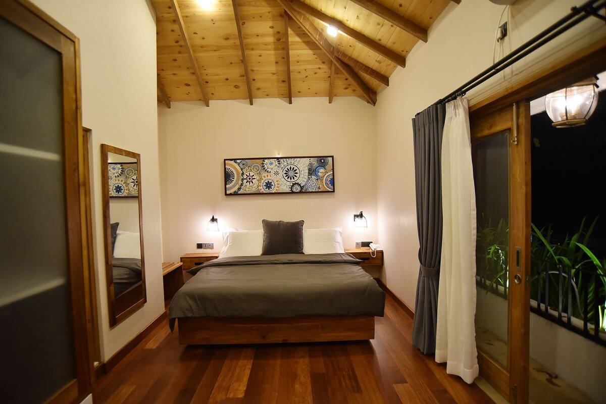 A bedroom with a wooden ceiling, a bed, two bedside lamps, a large window with curtains, and a wall-mounted artwork.