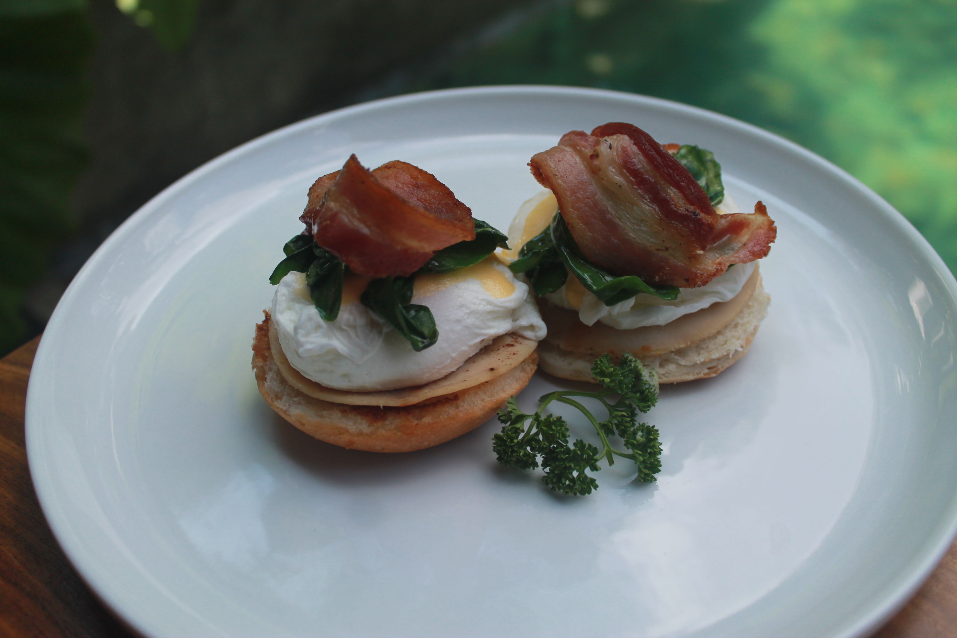 A plate with two eggs benedict sandwiches, garnished with bacon and parsley.