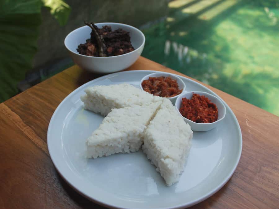 A plate of white rice with three sides, two small bowls of red sauce, and a bowl of dark-colored food on a wooden table.