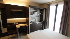 A hotel room with a bed, a desk with a chair, a built-in TV, a kitchenette with a microwave, and a window with curtains.