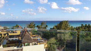 A balcony with a clear view of the ocean, palm trees, and a thatched-roof building. - New condo in best building Playa, 1 block from beach, amazing rooftop pool (Zazil-ha)