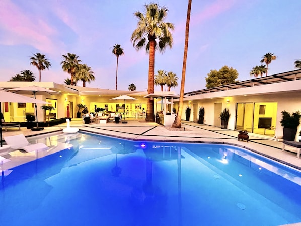 A swimming pool with lounge chairs and umbrellas, surrounded by palm trees and a modern building with a bar area.