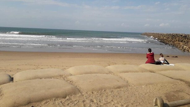 Una playa arenosa con un rompeolas rocoso y dos personas sentadas sobre la arena.