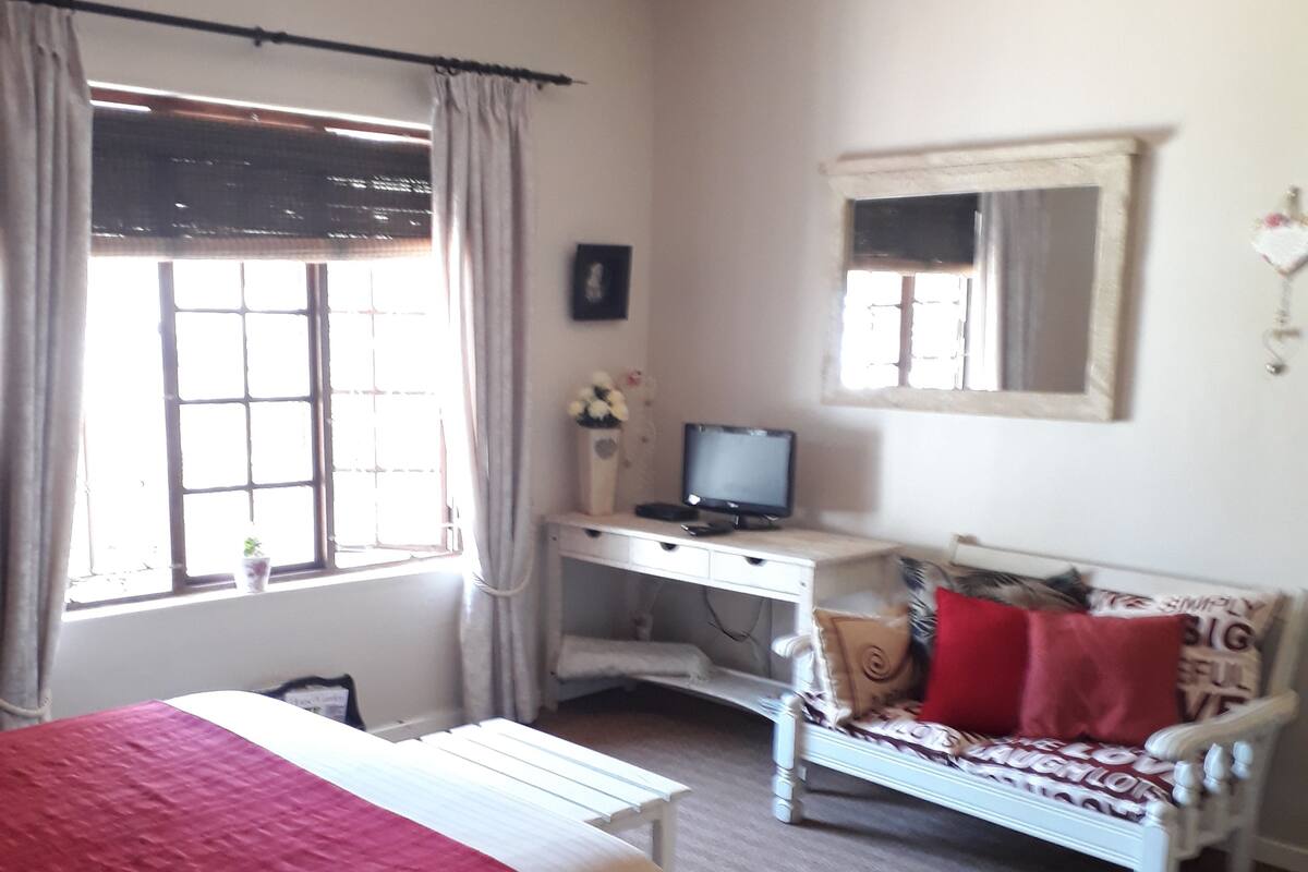 A bedroom with a bed, a desk, a chair, a sofa, a ceiling fan, a window, and a mirror.