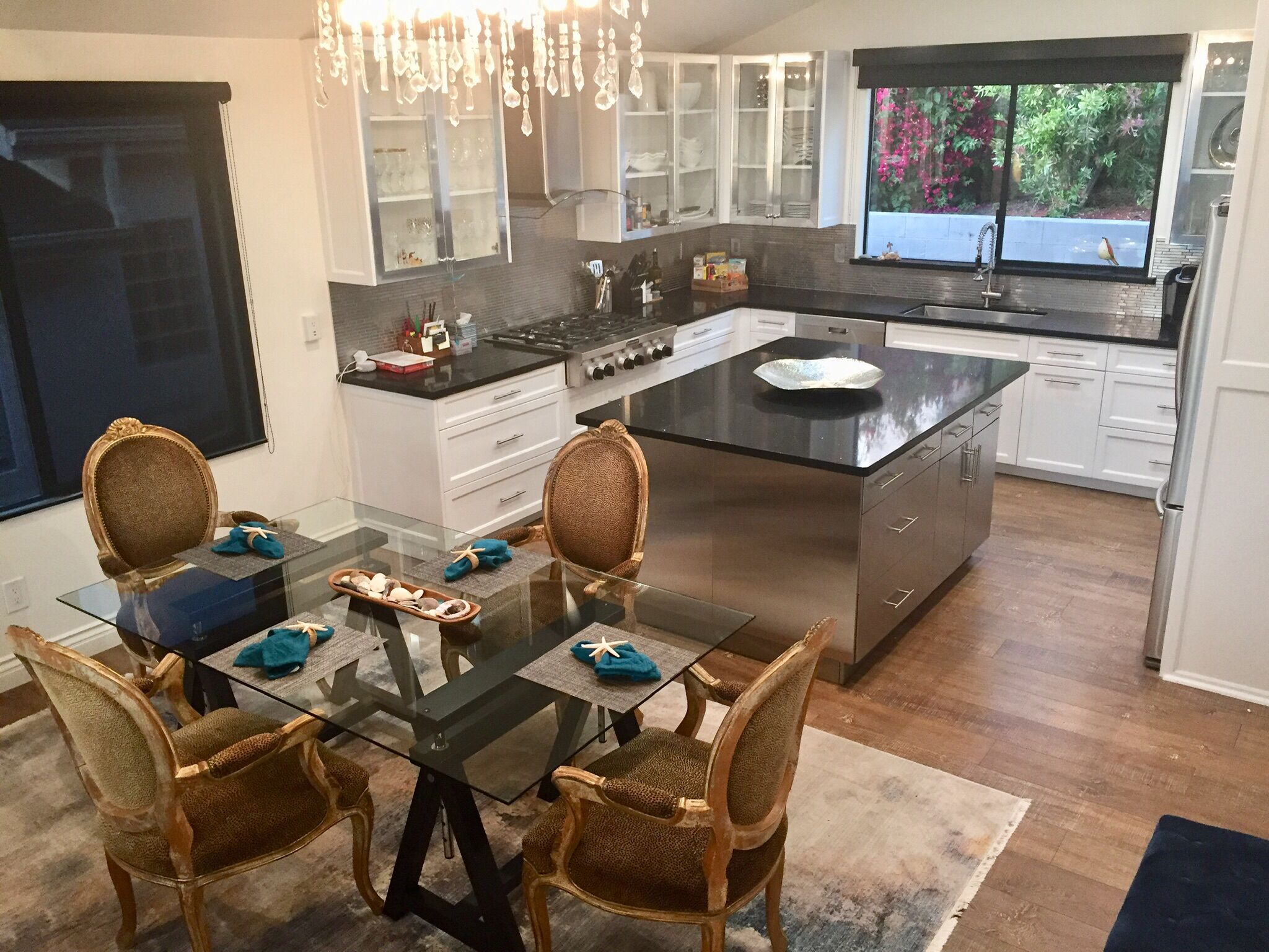 Una cocina moderna con una isla central, armarios blancos y un comedor con mesa de vidrio y sillas de mimbre.