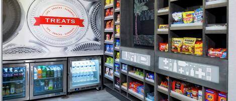 A snack and beverage area with refrigerated drinks, shelves stocked with snacks and treats, and a wall with a 'TREATS' sign.
