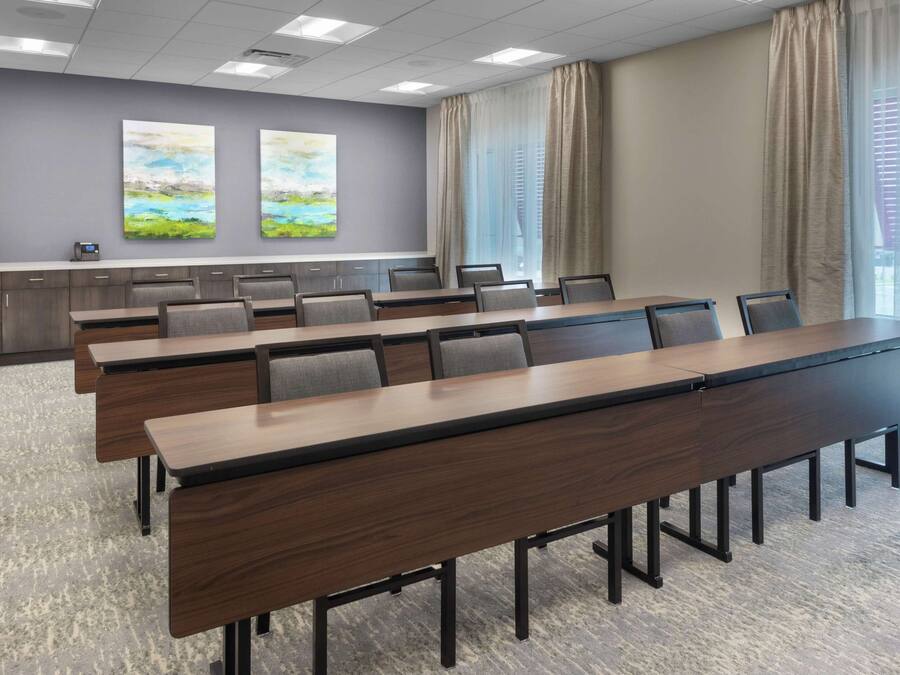 A conference room with long tables, chairs, and a grey carpet. There are two paintings on the wall and a window with curtains.