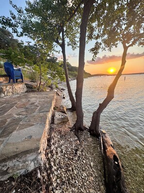 A riverside walkway with a stone bench, a blue plastic chair, and trees.