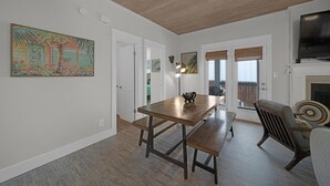 Un coin repas avec une table et des bancs en bois, un téléviseur placé au-dessus d’une cheminée, et une vue à travers une porte vitrée.