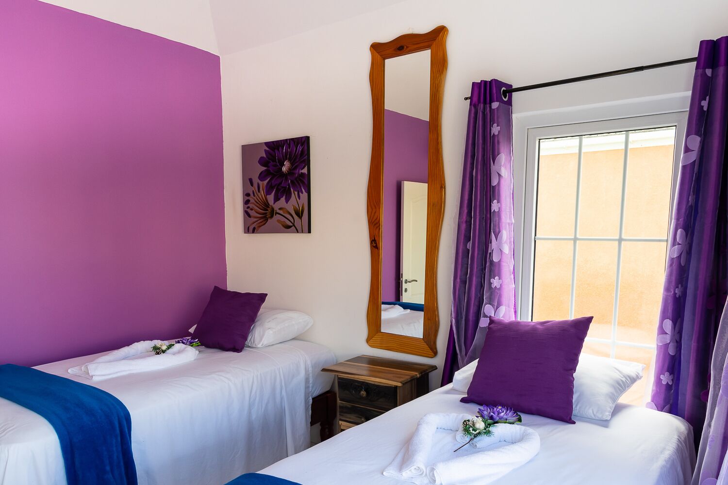 Un dormitorio con dos camas, cortinas moradas, un espejo grande y un cuadro floral en la pared.
