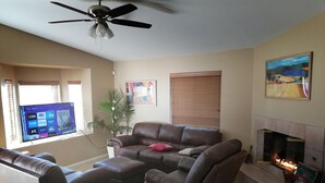 Un salon avec un canapé en cuir, une cheminée, un ventilateur de plafond et un téléviseur affichant la liste des chaînes.
