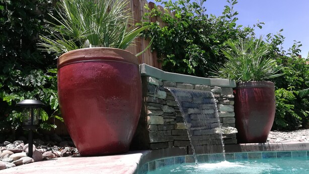 Une piscine agrémentée d’une cascade, entourée de grands bacs à fleurs rouges et d’un mur de pierre.